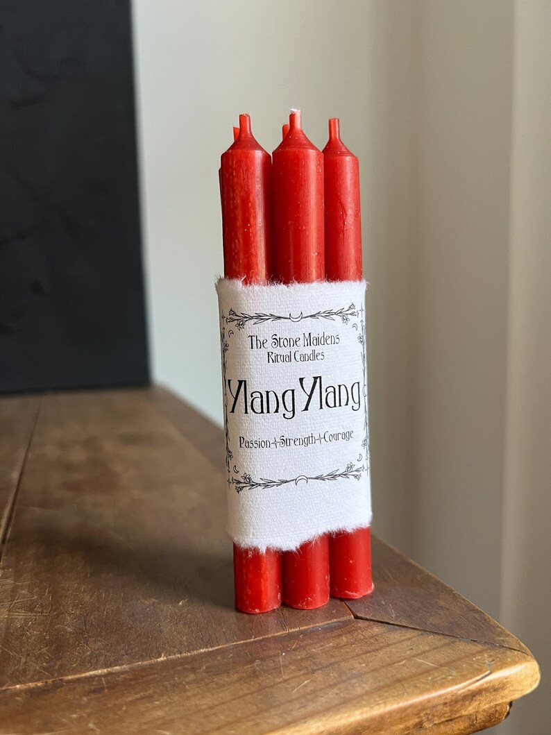 May include: A bundle of three red ritual candles. The candles are wrapped with a white label that reads "Ylang Ylang, Passion + Strength + Courage" and "The Stone Maidens Ritual Candles". The candles are on a wooden surface.