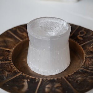 Puede incluir: Un portavelas cil&iacute;ndrico de selenita blanco transl&uacute;cido se encuentra dentro de un plato circular de color bronce. El plato presenta s&iacute;mbolos del zod&iacute;aco en relieve alrededor de su borde. El portavelas tiene una superficie rugosa y texturizada.