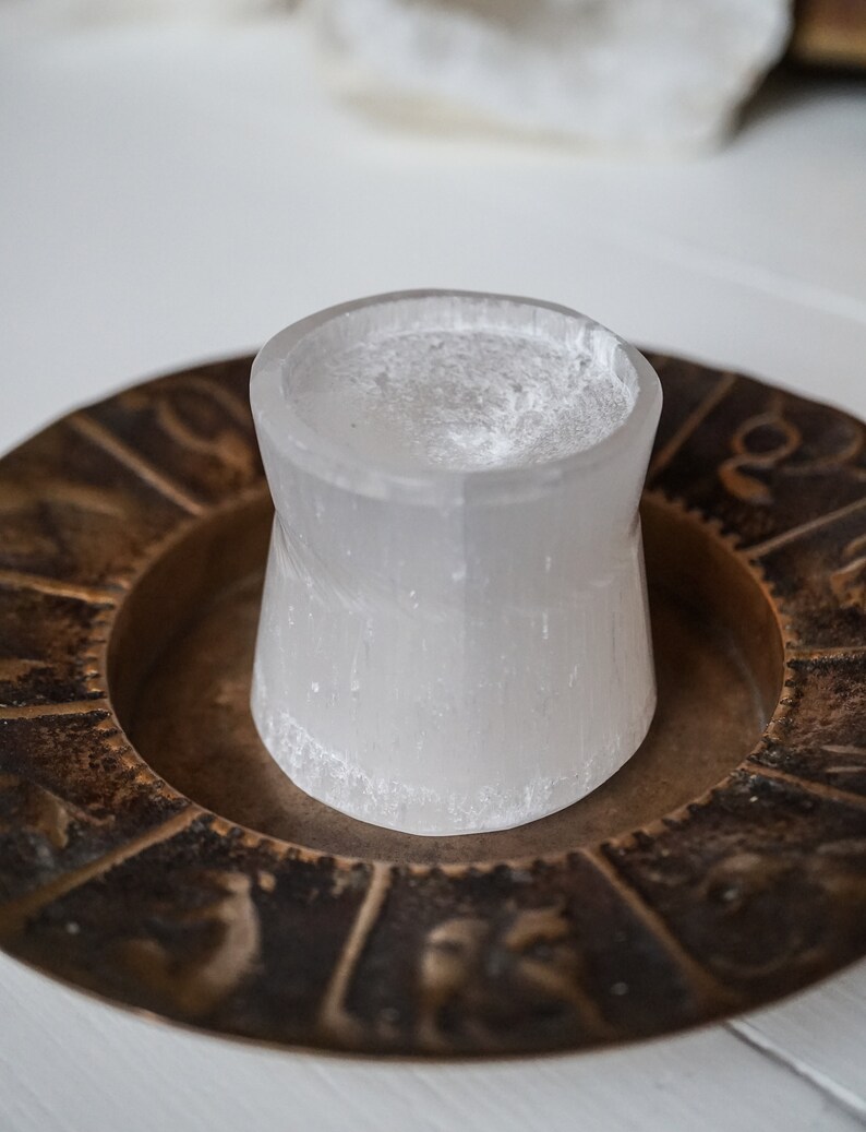 Puede incluir: Un portavelas cil&iacute;ndrico de selenita blanco transl&uacute;cido se encuentra dentro de un plato circular de color bronce. El plato presenta s&iacute;mbolos del zod&iacute;aco en relieve alrededor de su borde. El portavelas tiene una superficie rugosa y texturizada.
