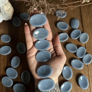 May include: A collection of smooth, oval-shaped blue celestite palm stones. The stones are arranged in a hand and on a wooden surface.