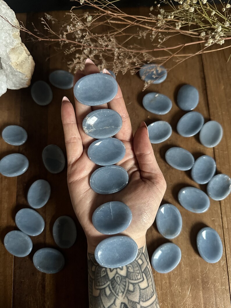 May include: A collection of smooth, oval-shaped blue celestite palm stones. The stones are arranged in a hand and on a wooden surface.