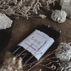 May include: A bundle of black candles wrapped in a white label with the word "Vetiver" printed on it. The label also includes the text "Protection-Banishing-Grounding". The candles are surrounded by dried flowers and decorative stones.