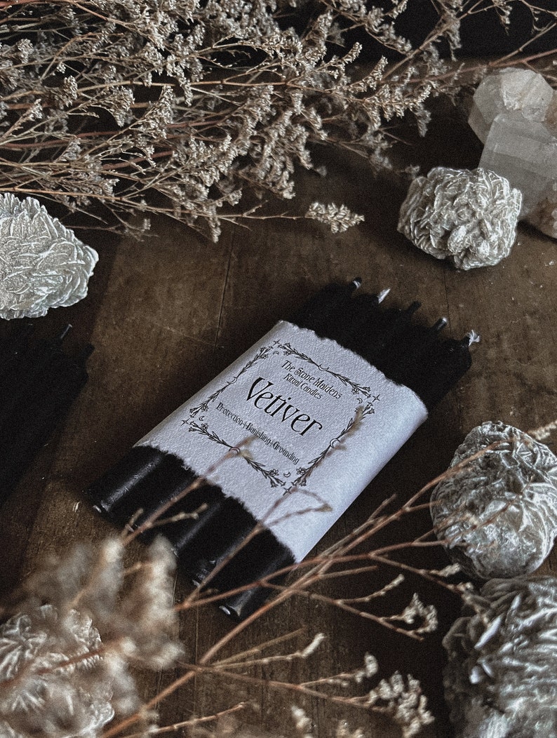 May include: A bundle of black candles wrapped in a white label with the word "Vetiver" printed on it. The label also includes the text "Protection-Banishing-Grounding". The candles are surrounded by dried flowers and decorative stones.