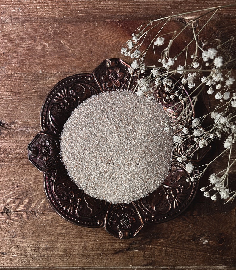 May include: A decorative metal tray with an ornate design, filled with white sand or small white beads. The tray is on a wooden surface with a sprig of white flowers.