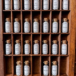 May include: A wooden display case filled with small glass bottles, each labeled with a different gemstone name. The bottles have cork stoppers and white labels with black text. The gemstones include Amethyst, Quartz, and Tourmaline.