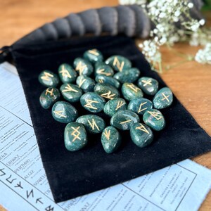 May include: A set of 25 green agate rune stones with gold lettering on each stone. The stones are in a black velvet pouch.