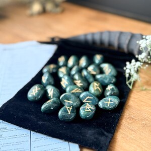 May include: A set of 25 green agate rune stones with gold lettering on a black velvet pouch. The runes are arranged in a semi-circle on the pouch.