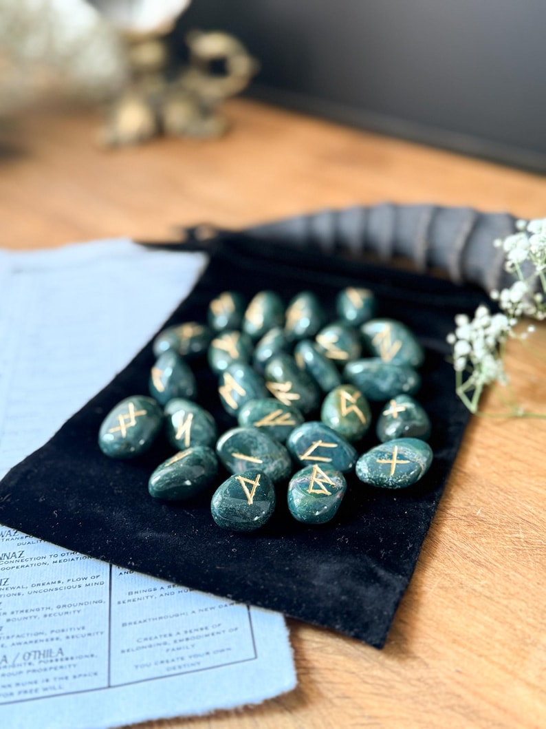 May include: A set of 25 green agate rune stones with gold lettering on a black velvet pouch. The runes are arranged in a semi-circle on the pouch.