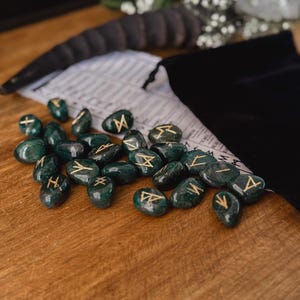 May include: A set of green gemstone runes with gold lettering. The runes are scattered on a wooden surface.