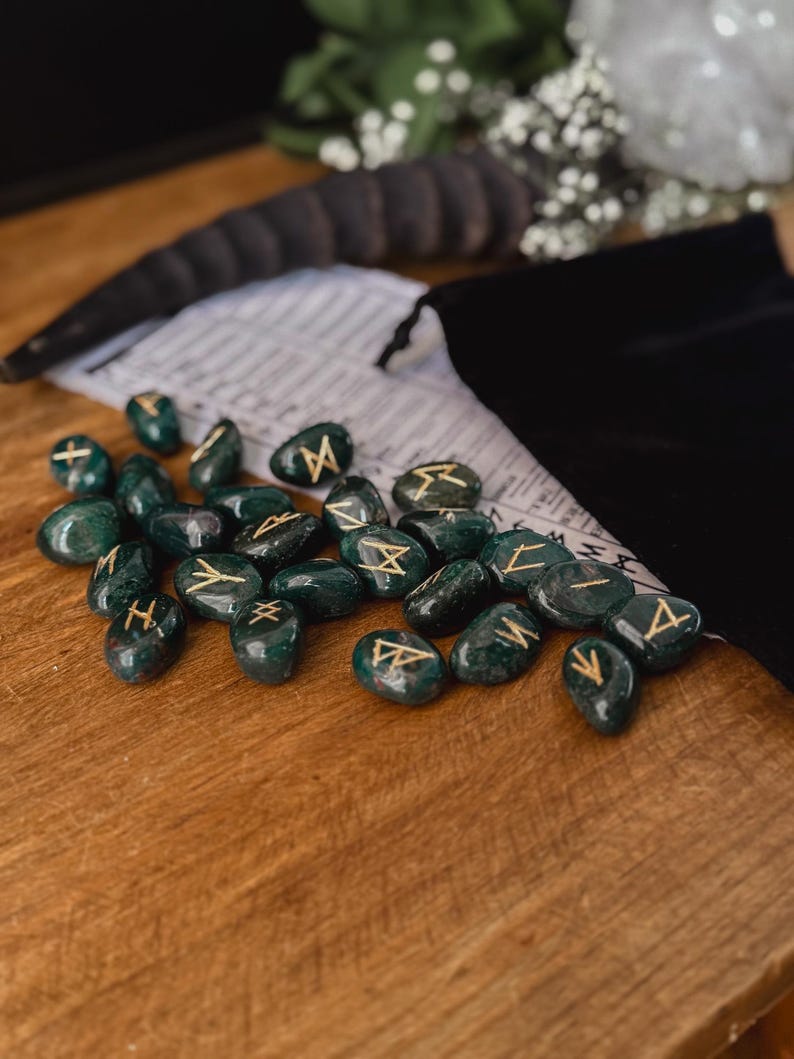 May include: A set of green gemstone runes with gold lettering. The runes are scattered on a wooden surface.