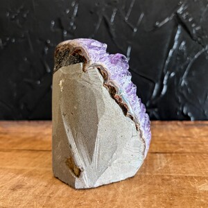 May include: A raw amethyst geode specimen featuring a cluster of deep purple, crystalline points emerging from a jagged, grey stone base. The mineral formation displays a natural, uneven texture with a distinct boundary between the rough exterior rock and the vibrant, translucent violet crystals that catch the light.