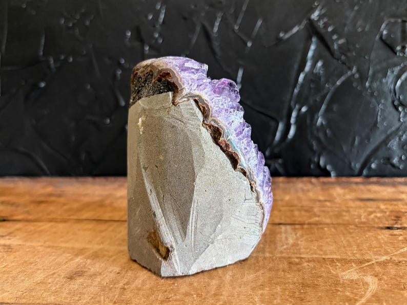 May include: A raw amethyst geode specimen featuring a cluster of deep purple, crystalline points emerging from a jagged, grey stone base. The mineral formation displays a natural, uneven texture with a distinct boundary between the rough exterior rock and the vibrant, translucent violet crystals that catch the light.