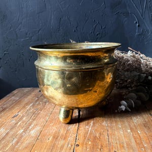 May include: A polished brass bowl with a wide, flared rim and a rounded body. The bowl is supported by three small, rounded feet at the base, featuring a smooth, reflective metallic surface with subtle signs of natural aging and patina.