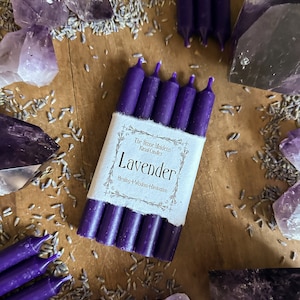 Puede incluir: Seis velas de color púrpura envueltas en papel blanco con el texto "The Stone Maidens Ritual Candles Lavender Healing + Wisdom + Intuition" impreso en el papel. Las velas están rodeadas de capullos de lavanda y cristales de amatista.
