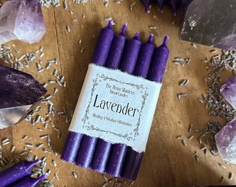 May include: Six purple candle sticks wrapped in white paper with the text "The Stone Maidens Ritual Candles Lavender Healing + Wisdom + Intuition" printed on the paper. The candles are surrounded by lavender buds and amethyst crystals.