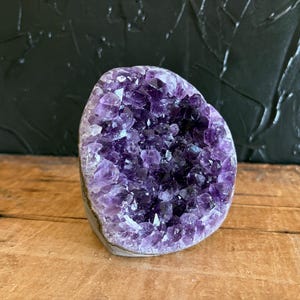 May include: A raw amethyst geode featuring a cluster of deep purple, multifaceted crystal points. The specimen has a rounded, irregular shape with a natural, rough exterior edge and a hollow center filled with vibrant, translucent violet crystals.