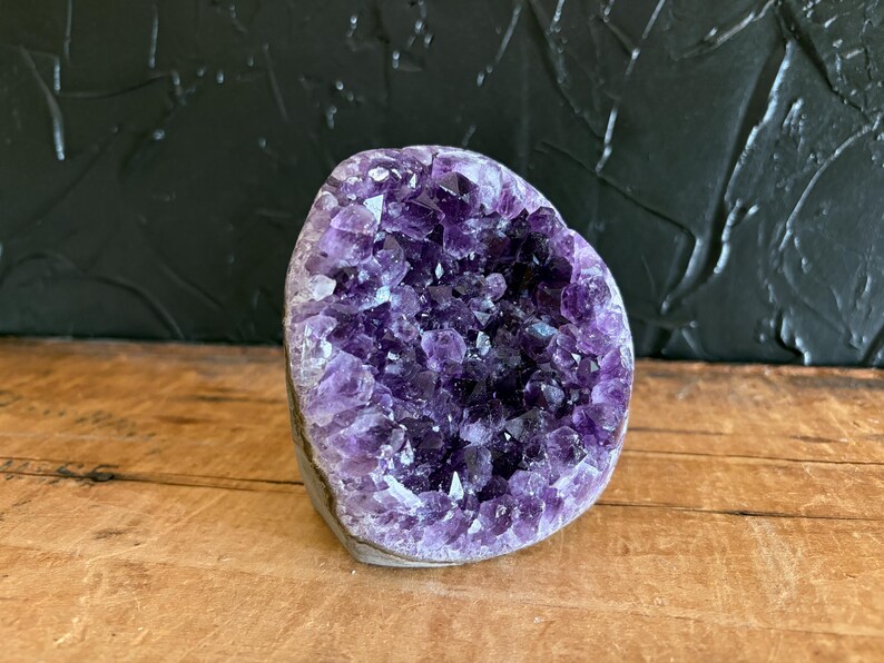 May include: A raw amethyst geode featuring a cluster of deep purple, multifaceted crystal points. The specimen has a rounded, irregular shape with a natural, rough exterior edge and a hollow center filled with vibrant, translucent violet crystals.