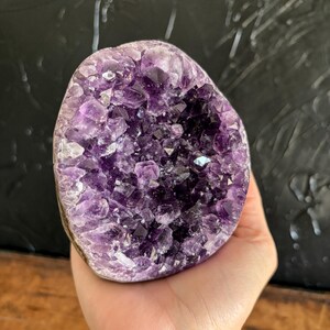 May include: An amethyst geode featuring a cluster of deep purple, multifaceted crystal points. The interior cavity is densely packed with jagged, translucent violet formations that transition into darker, saturated purple tones toward the center.