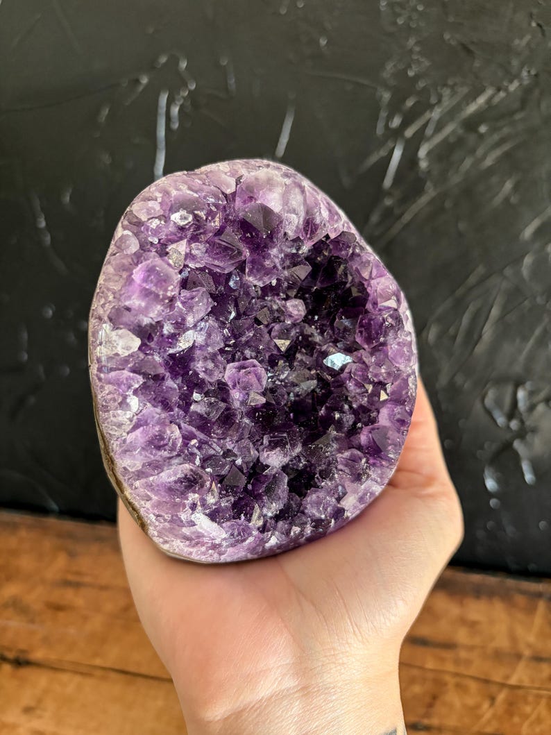 May include: An amethyst geode featuring a cluster of deep purple, multifaceted crystal points. The interior cavity is densely packed with jagged, translucent violet formations that transition into darker, saturated purple tones toward the center.