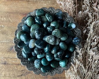 May include: A bowl filled with green moss agate tumbled stones. The stones are smooth and polished, with a variety of green shades and patterns.