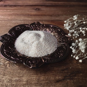 May include: A small, ornate, copper-colored dish with a pile of white sand in the center. The dish is on a wooden surface with a sprig of white flowers to the right.
