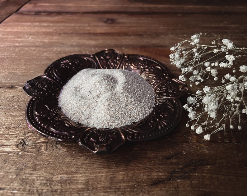 May include: A small, ornate, copper-colored dish with a pile of white sand in the center. The dish is on a wooden surface with a sprig of white flowers to the right.