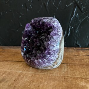 May include: A natural amethyst geode featuring a hollow center filled with deep purple, crystalline points. The exterior of the stone displays a rough, earthy texture with shades of grey and brown, contrasting with the vibrant violet interior.