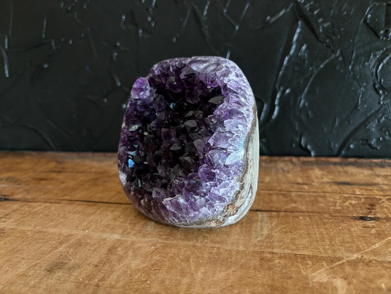 May include: A natural amethyst geode featuring a hollow center filled with deep purple, crystalline points. The exterior of the stone displays a rough, earthy texture with shades of grey and brown, contrasting with the vibrant violet interior.