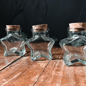 May include: Three clear, star-shaped glass bottles with cork stoppers are arranged on a wooden surface. The bottles are empty and have a vintage appearance. The dark background provides a contrast to the bottles.
