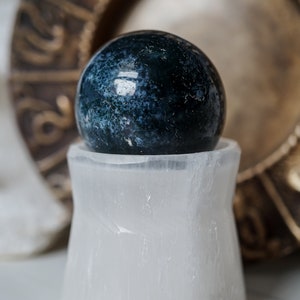 Puede incluir: Una esfera de piedra preciosa azul oscuro y verde descansa sobre un soporte de cristal de selenita blanco. La esfera tiene un patr&oacute;n moteado. En el fondo hay un plato decorativo de color dorado.
