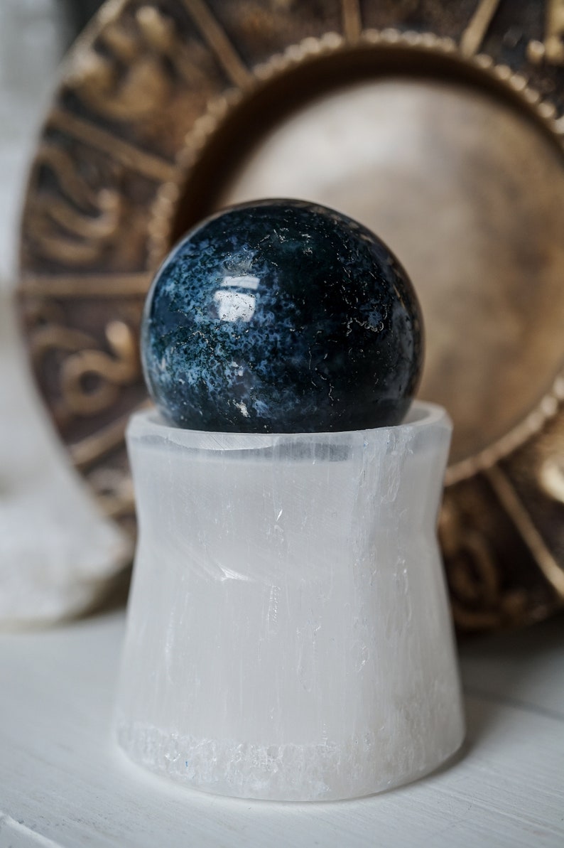 Puede incluir: Una esfera de piedra preciosa azul oscuro y verde descansa sobre un soporte de cristal de selenita blanco. La esfera tiene un patr&oacute;n moteado. En el fondo hay un plato decorativo de color dorado.