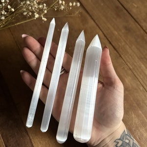 May include: Four white selenite wands of varying lengths, with pointed tips, are held in a hand. The wands have a striated texture.