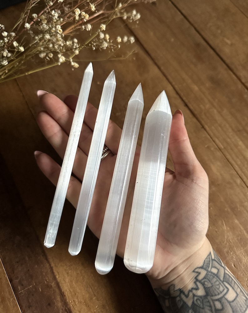 May include: Four white selenite wands of varying lengths, with pointed tips, are held in a hand. The wands have a striated texture.