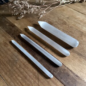 May include: Five selenite wands of varying lengths, lying on a wooden surface. The wands are smooth and white.