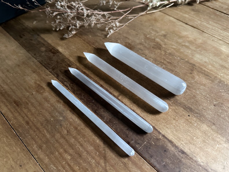 May include: Five selenite wands of varying lengths, lying on a wooden surface. The wands are smooth and white.