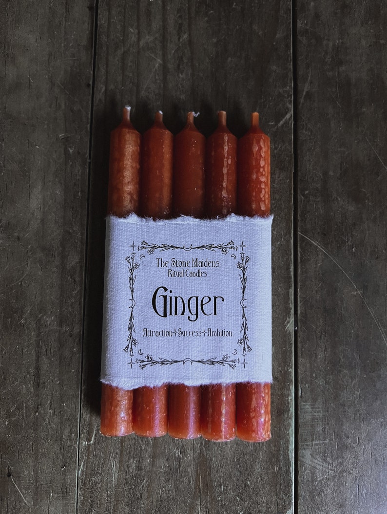 K&ouml;nnte beinhalten: Ein Set mit sechs orangefarbenen Ingwer-Ritualkerzen, verpackt in wei&szlig;em Stoff mit einem Etikett mit der Aufschrift "The Stone Maidens Ritual Candles Ginger Attraction + Success + Ambition".