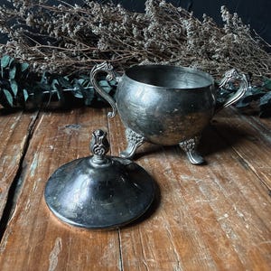 May include: A silver-toned metal sugar bowl with two ornate handles and three decorative feet, accompanied by a matching circular lid featuring a central finial. The bowl has a rounded body and a tarnished, metallic finish throughout.