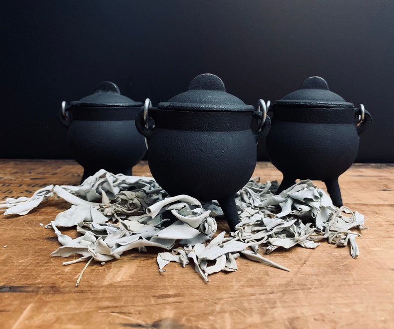 May include: Three small black cast iron cauldrons with silver handles sitting on a bed of white sage.