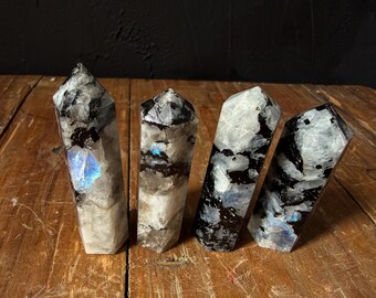 May include: Four polished crystal towers with a mix of black, white, and grey patterns. Some crystals have a blue iridescent flash. The crystals are displayed on a wooden surface.