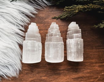 May include: Three white selenite tower crystals with a layered design, displayed on a dark wood surface. The crystals have a tiered design. A fluffy white material and greenery are in the background.