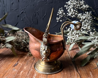 May include: A copper coal scuttle with a brass base and a swing handle. The handle features a decorative lion head mount on each side and a blue and white floral porcelain grip. The vessel has a wide, open top and a rounded, polished body.