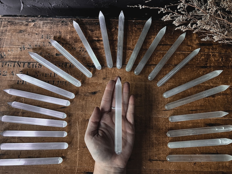 May include: A collection of selenite wands, also known as selenite sticks, arranged in a semi-circle on a wooden surface. The wands are white and translucent, with a smooth, polished finish. One wand is held in a hand in the centre of the image.