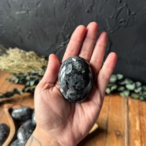 May include: A dark grey and black speckled, oval-shaped stone is held in an open hand. Other similar stones are visible in a wooden bowl. The background features a dark, textured surface. The stone has a polished finish.
