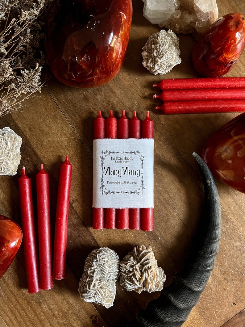 May include: A collection of red ritual candles, crystals, and a horn on a wooden surface. The candles are packaged with a label that reads "Ylang Ylang Passion + Strength + Courage". Various crystals and stones are arranged around the candles.