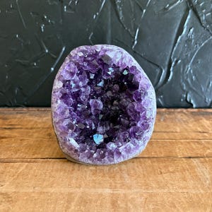 May include: A natural amethyst geode featuring a cluster of deep purple, crystalline points within a rounded, stone-like exterior. The interior cavity is densely packed with jagged, translucent violet crystals that catch the light, creating a textured, multifaceted appearance.