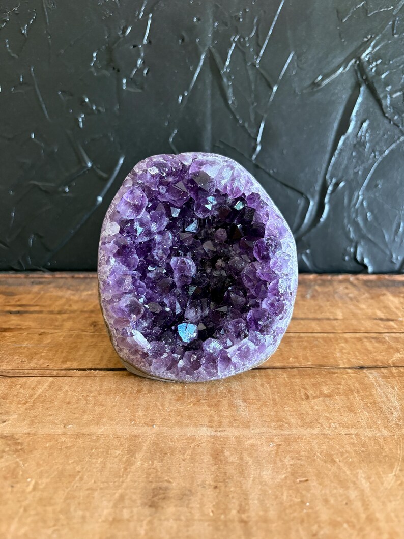 May include: A natural amethyst geode featuring a cluster of deep purple, crystalline points within a rounded, stone-like exterior. The interior cavity is densely packed with jagged, translucent violet crystals that catch the light, creating a textured, multifaceted appearance.