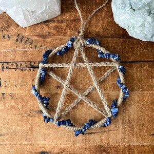 May include: A five-pointed star, or pentagram, made of twine with blue lapis lazuli stones around the perimeter. The star is hanging from a twine loop.