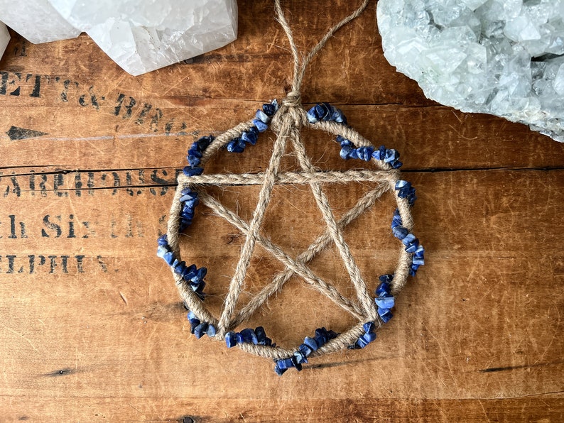 May include: A five-pointed star, or pentagram, made of twine with blue lapis lazuli stones around the perimeter. The star is hanging from a twine loop.