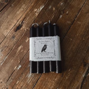 May include: A bundle of black ritual candles wrapped with a white label. The label features a raven illustration and text that reads "The Stone Maidens Ritual Candles" and "Protection, Banishing, Grounding, Obsidian, Dark."