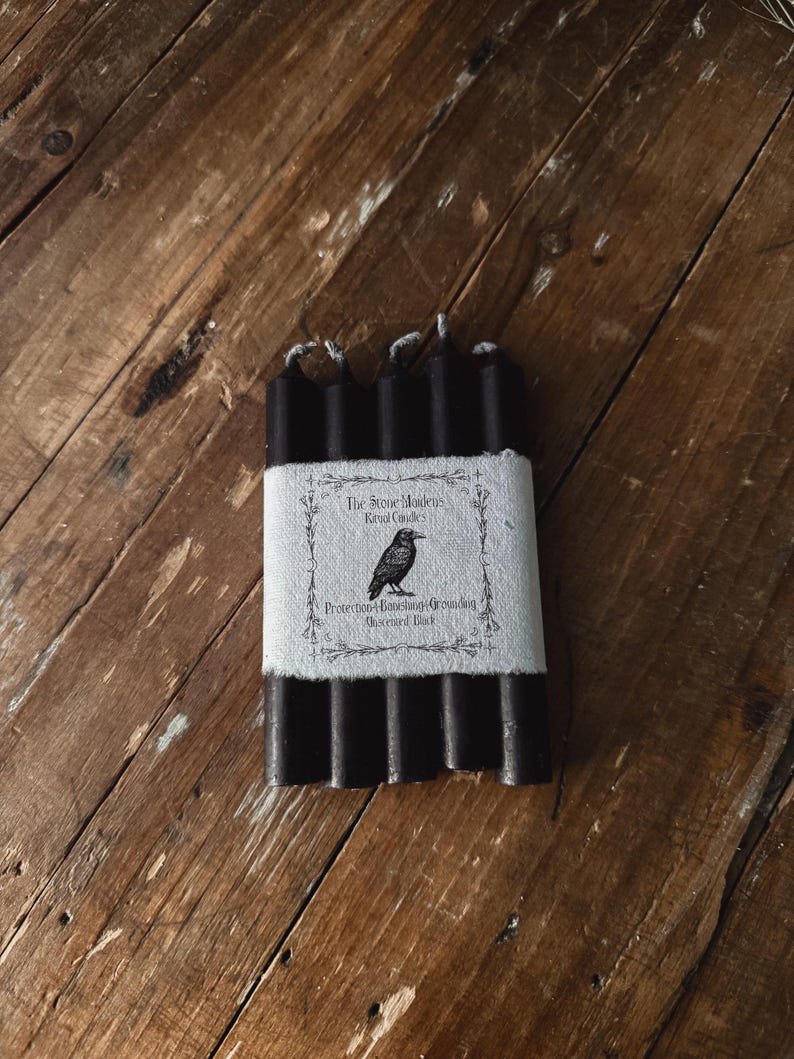 May include: A bundle of black ritual candles wrapped with a white label. The label features a raven illustration and text that reads "The Stone Maidens Ritual Candles" and "Protection, Banishing, Grounding, Obsidian, Dark."